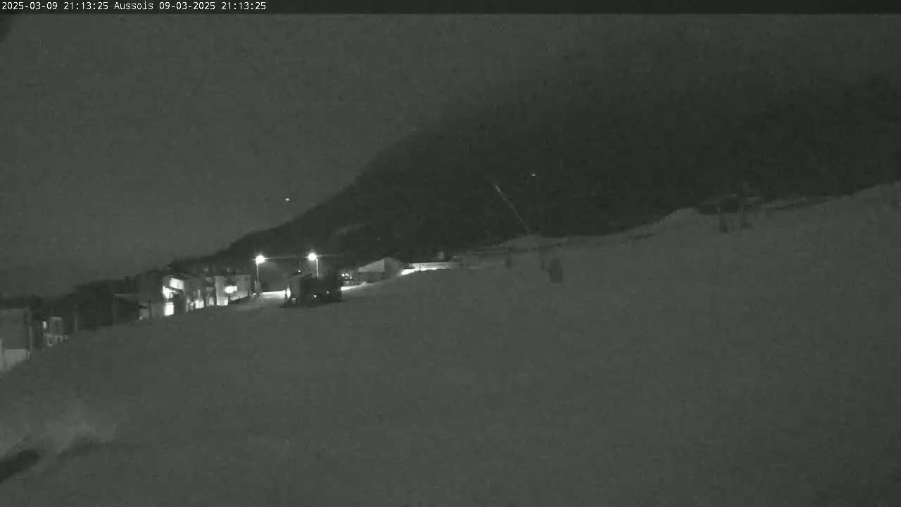 A snow-covered nighttime scene shows a few buildings and a vehicle near a dark mountain under a cloudy sky.
