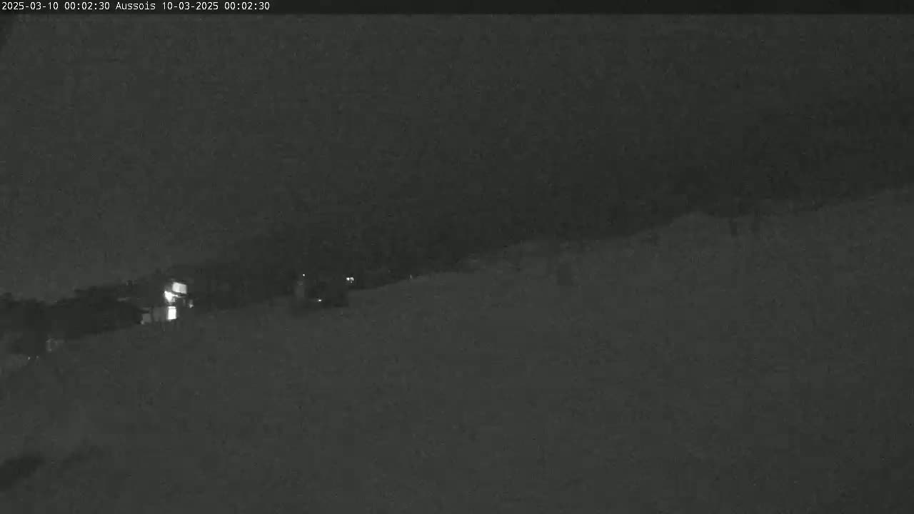A nighttime, snowy scene shows a dimly lit area with faint structures and minimal activity.