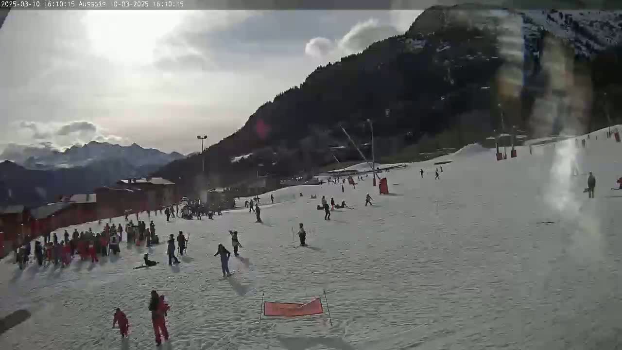 A snow-covered ski slope with skiers and snowboarders, some buildings, and a mountain range in the background under a partly cloudy sky.