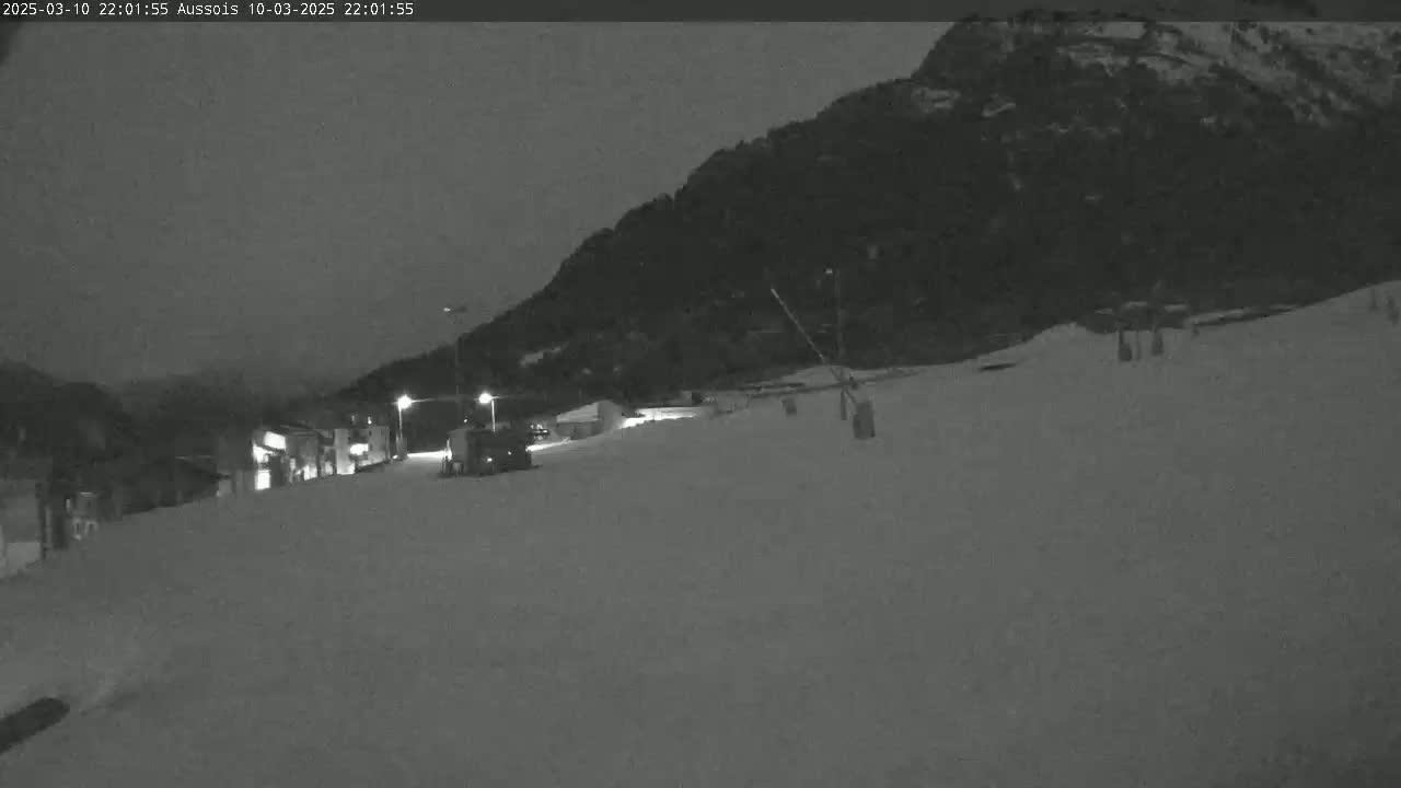 A snow-covered slope at night is illuminated by artificial lights, with a snowcat visible near some buildings and a dark mountain in the background.