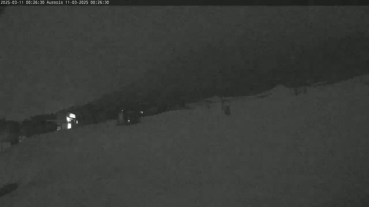 A nighttime, low-light image shows a snow-covered landscape with a few faint lights in the distance.