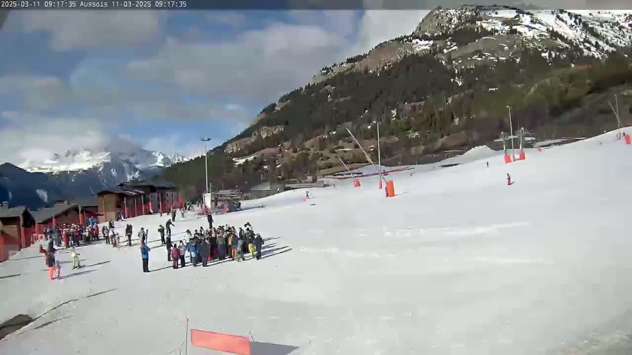 A snowy ski slope with several skiers and a group of people standing near buildings in the background under a partly cloudy sky.