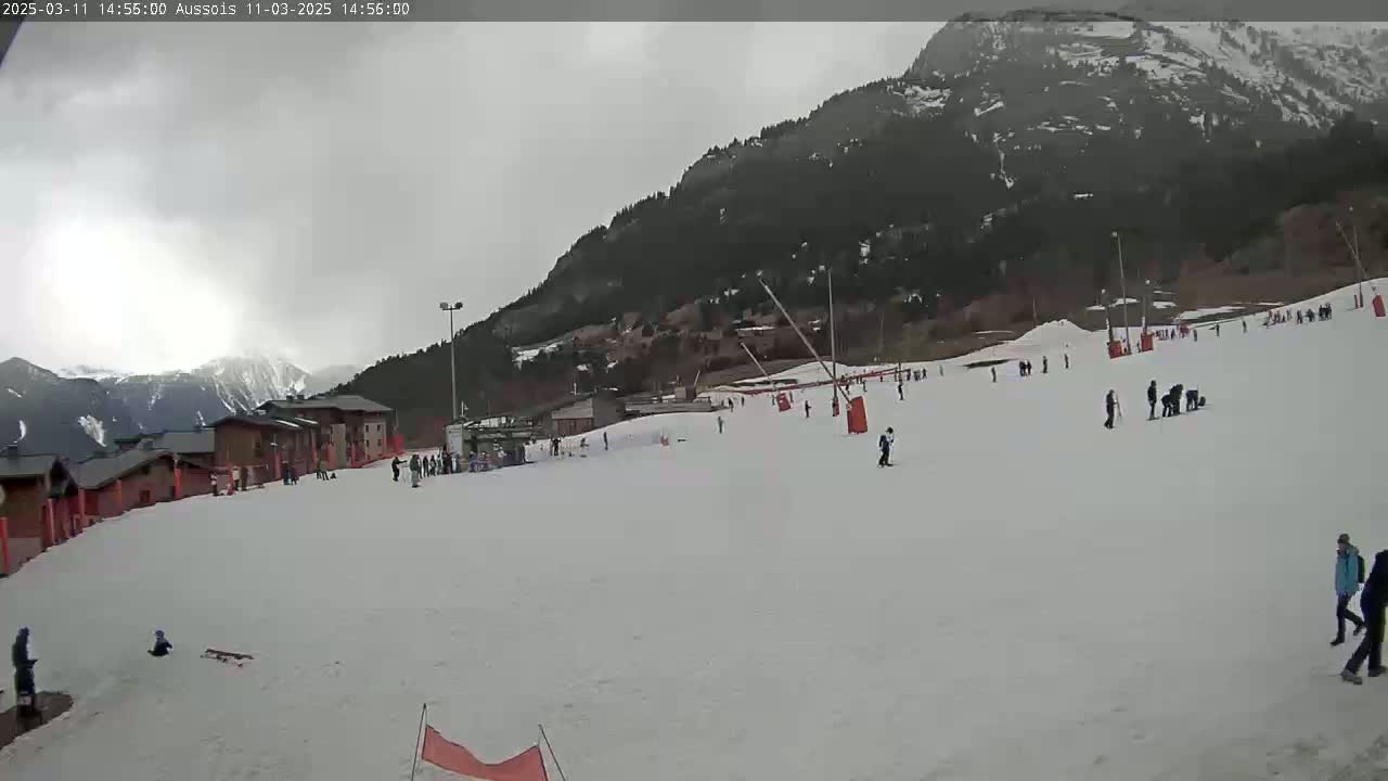 A snow-covered ski slope with several skiers and snowboarders, buildings, and a mountain in the background under an overcast sky.