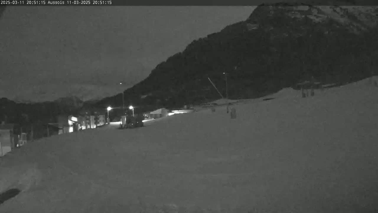 A nighttime, snow-covered scene shows a mountain in the background and a few lights illuminating a flat, snow-covered area in the foreground with some small vehicles.