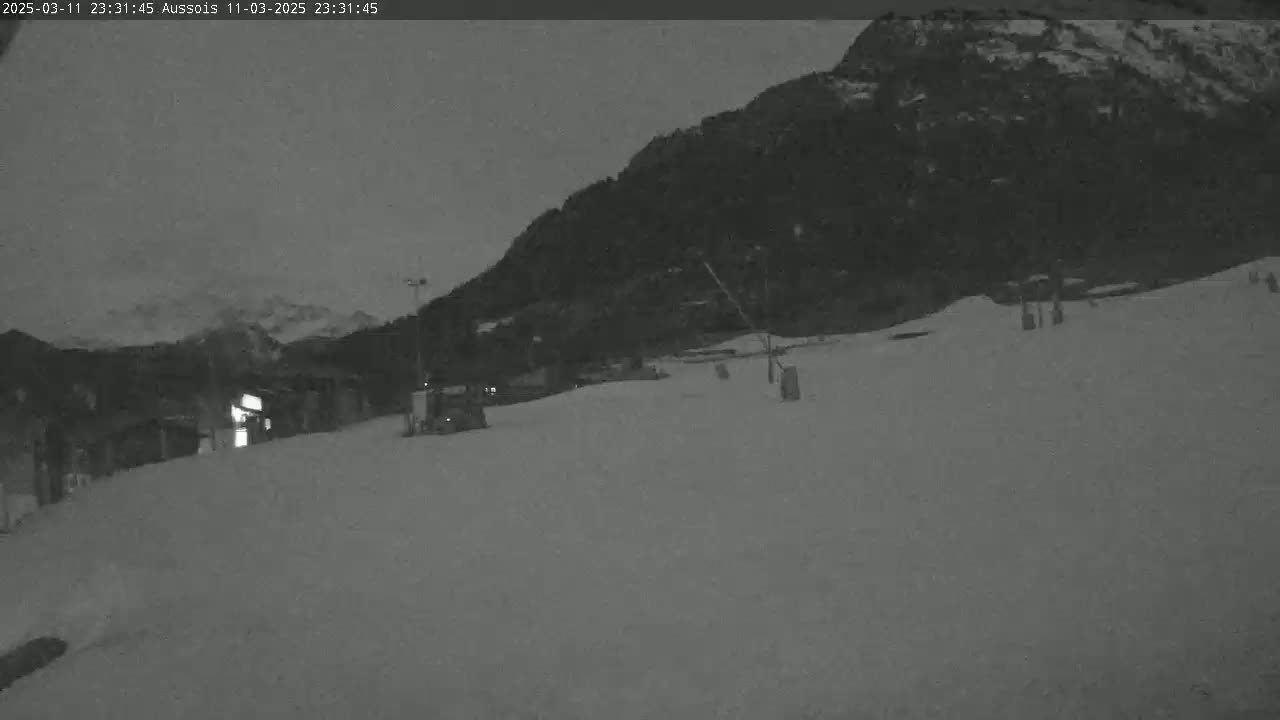 A snow-covered slope at night, with a dark mountain in the background under a dim, overcast sky.