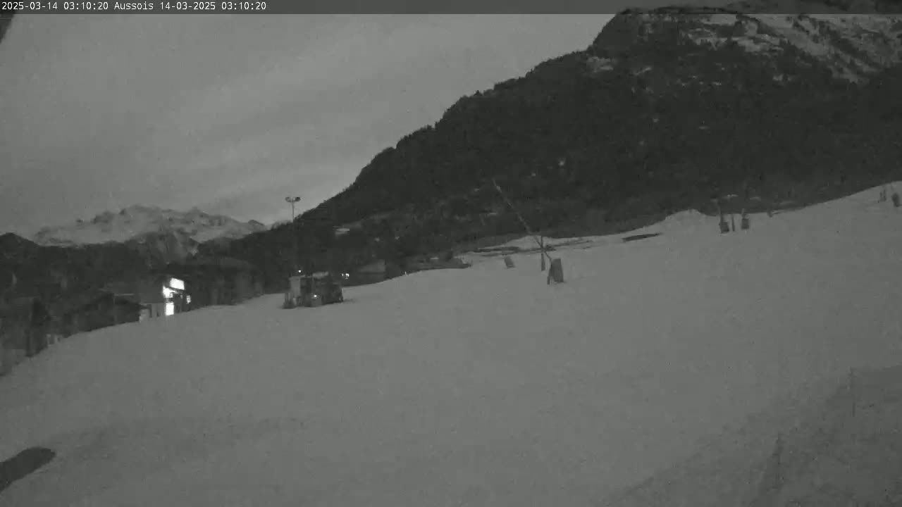 A snow-covered slope at twilight, with a dark mountain in the background and a few structures visible in the distance.