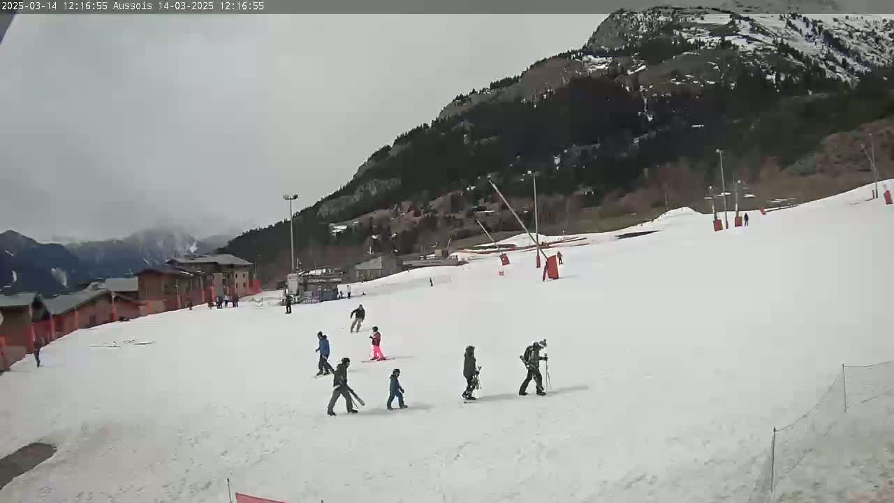 A snowy ski slope with several skiers and snowboarders, under an overcast sky, is nestled against a backdrop of mountains covered in trees.