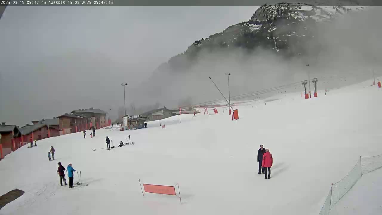 A snowy ski slope with several people scattered across it, under foggy, overcast conditions, with a mountain shrouded in fog in the background.
