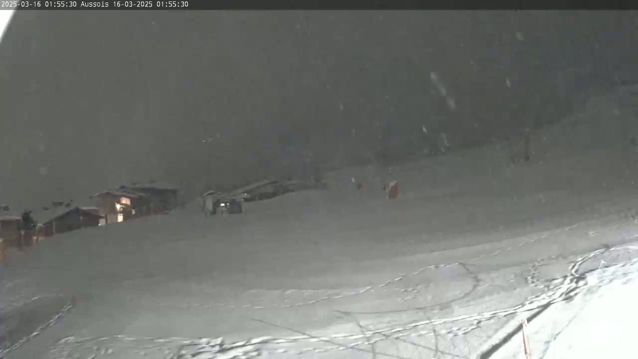 A snowy, nighttime scene shows a snow-covered slope with a few buildings visible in the distance under heavy snowfall.