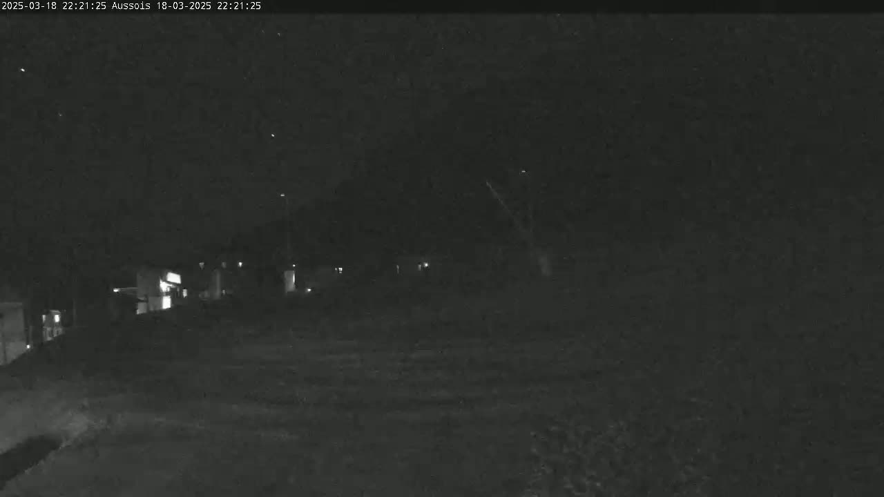 A nighttime, low-light image shows a dark, possibly rural area with a few dimly lit buildings in the distance.
