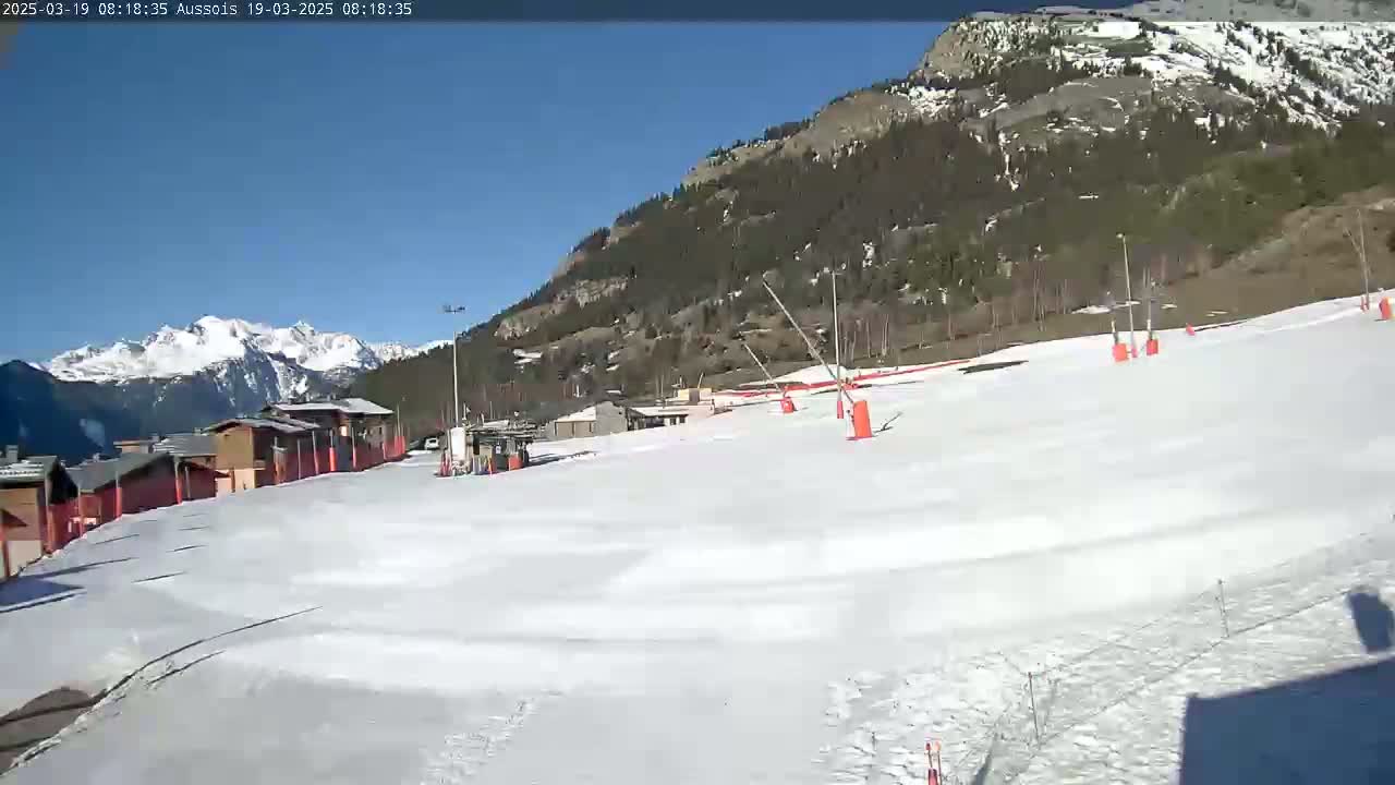 A snow-covered ski slope with buildings and a mountain range in the background under a sunny sky.