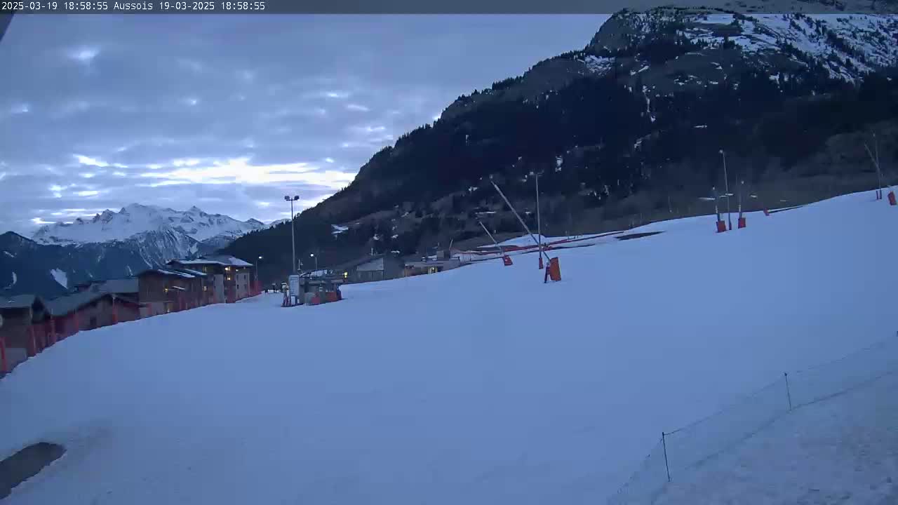 A snow-covered ski slope with buildings and a mountain in the background under a twilight sky.