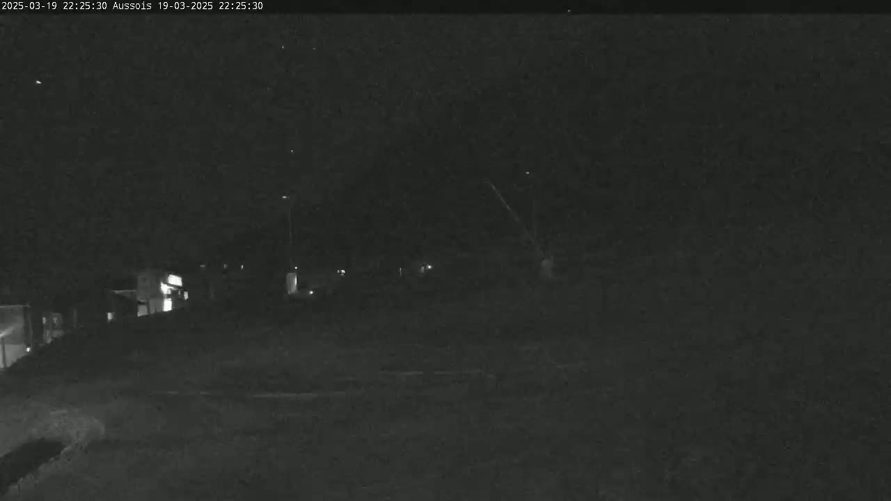 A nighttime, low-light view shows dimly lit buildings and a dark, possibly snowy, ground.
