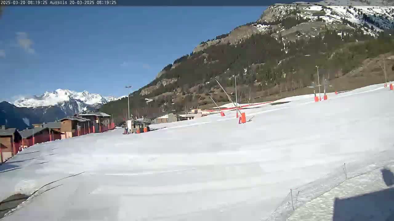 A snow-covered ski slope with several orange lift poles and a few buildings in the background under a clear blue sky.