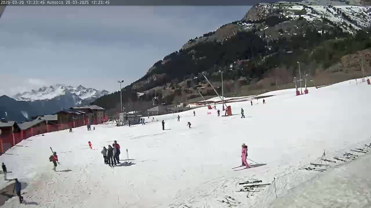 A snowy ski slope with several skiers and snowboarders is nestled against a mountainside under a mostly clear sky.