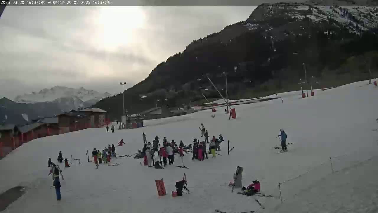 A snowy ski slope with several people, some skiing and others standing in groups, is situated below a mountain under a mostly cloudy sky.