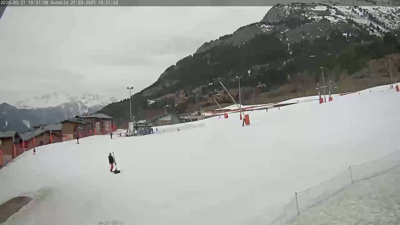 A snowy ski slope with a few skiers and buildings in the background under an overcast sky.