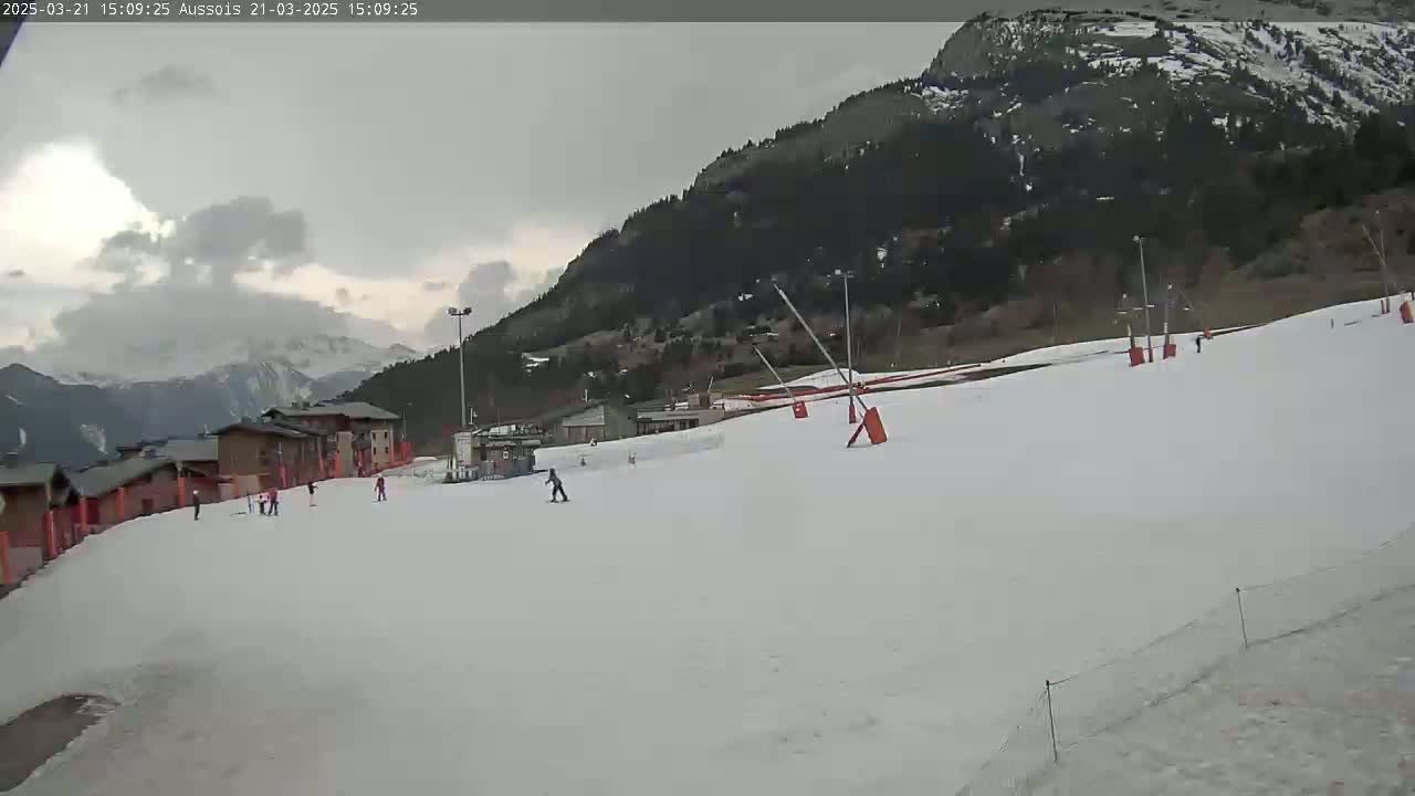 A snowy ski slope with several skiers and snowboarders visible, under an overcast sky, is situated in a mountainous region.
