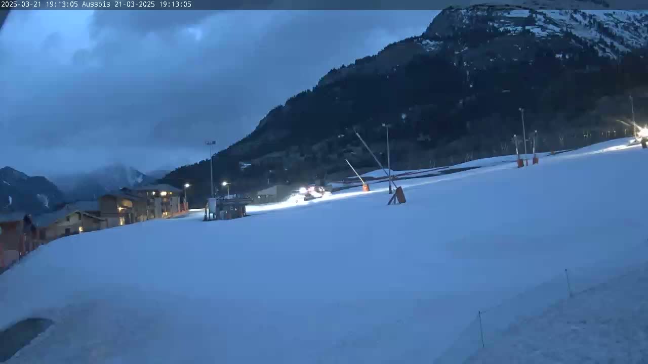 A snow-covered ski slope at dusk or night, with buildings and a mountain in the background under a cloudy sky.
