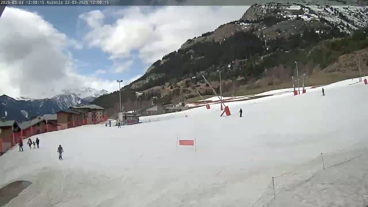 A mostly clear, partly cloudy day reveals a snow-covered ski slope with several skiers and buildings in the background, nestled against a mountainside covered with dark evergreen trees.