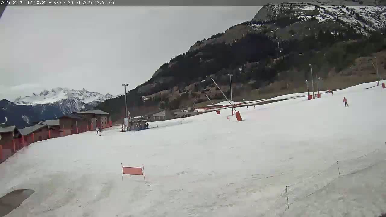 A snow-covered ski slope with several skiers, a few buildings, and mountains in the background under an overcast sky.