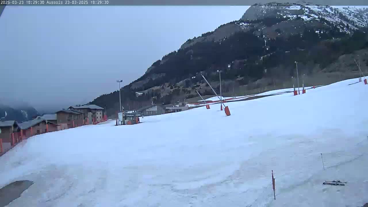 A snow-covered ski slope with a few buildings and ski lifts in the foreground, and a mountain in the background under an overcast sky.