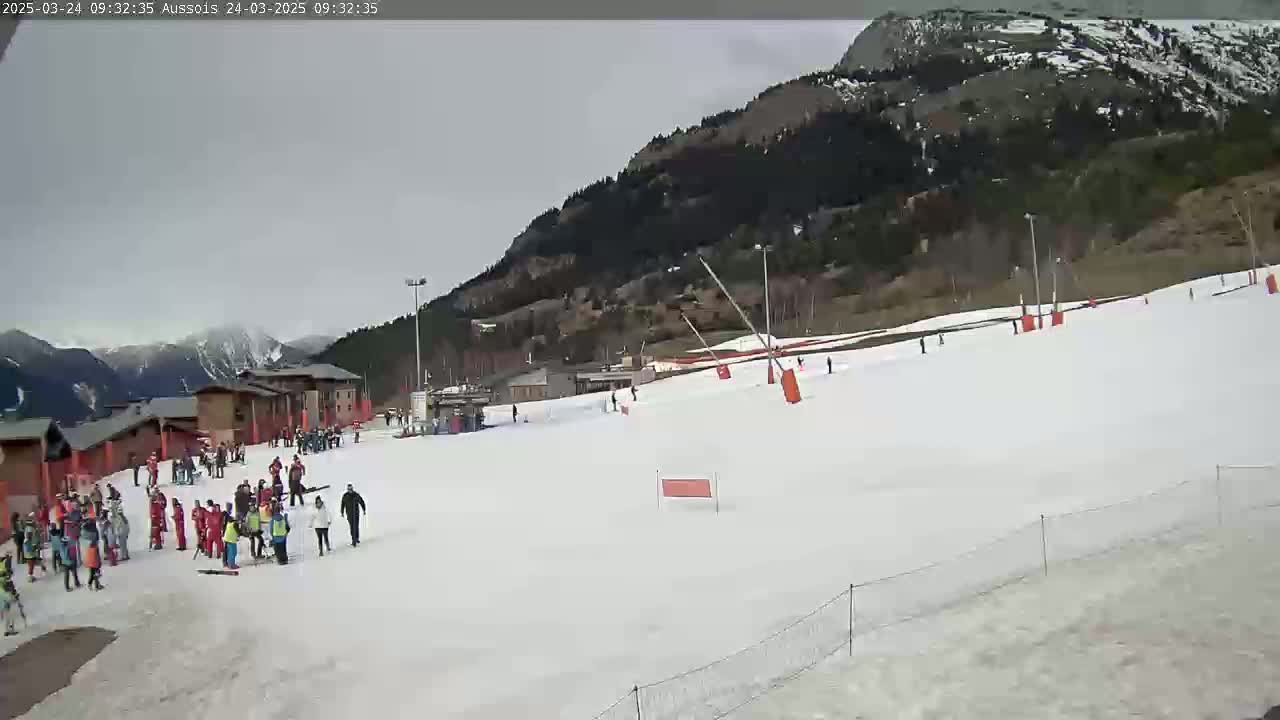 A snow-covered ski slope with several people skiing and standing around, buildings, and mountains in the background under an overcast sky.