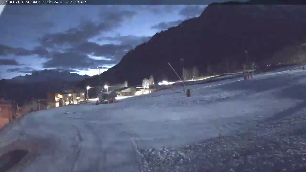 A snow-covered slope at dusk, illuminated by lights, with a mountain range in the background under a cloudy sky.