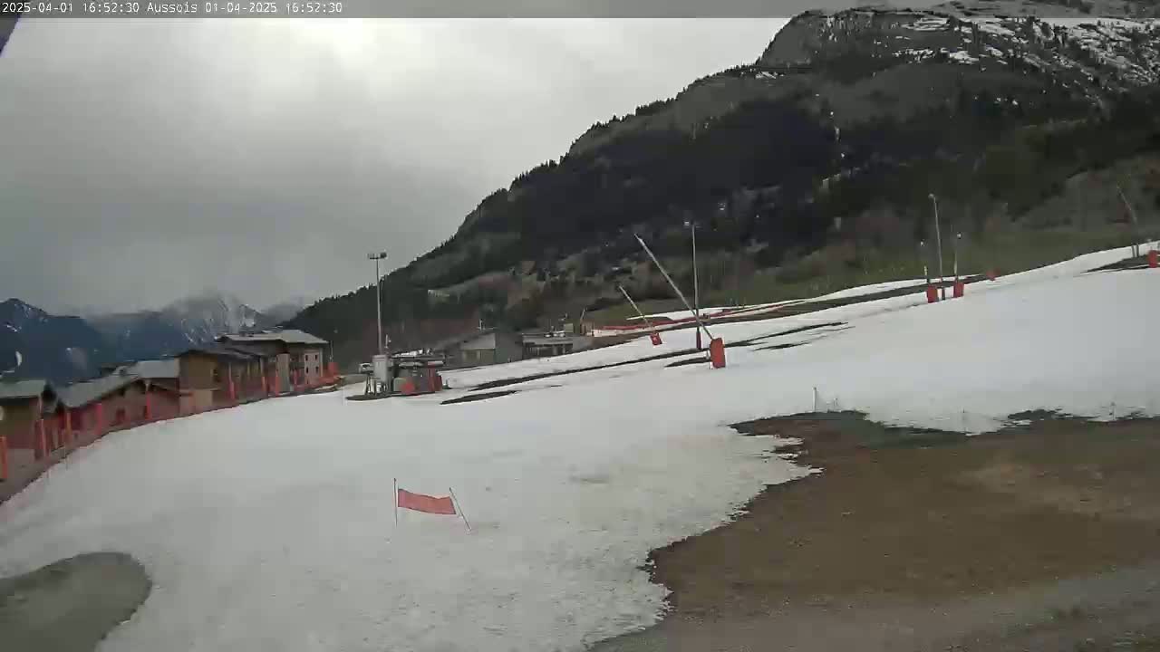 A snow-covered ski slope is situated below a mountain range under an overcast sky.