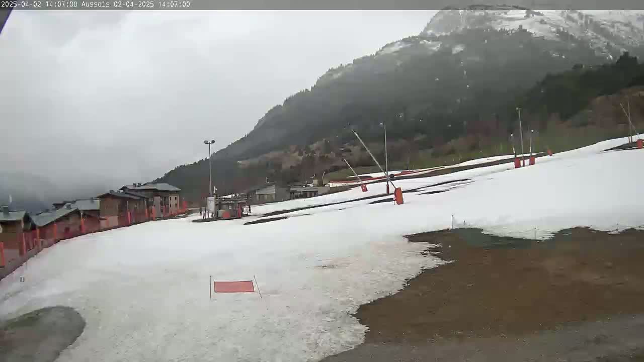 A snow-covered ski slope sits below a mountain range under an overcast sky.