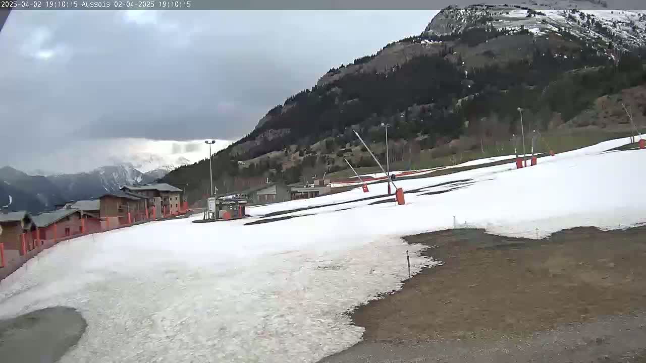 A partially snow-covered ski slope sits below a mountain under a cloudy sky.