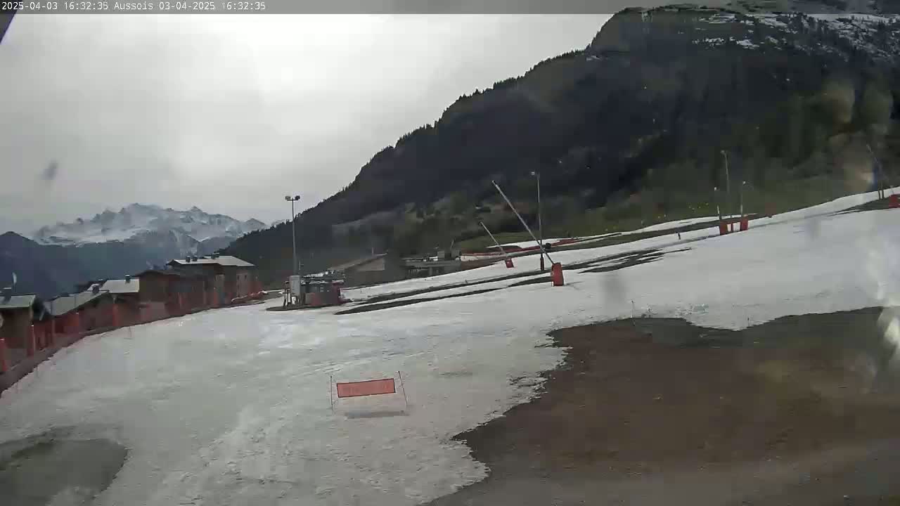 A mostly cloudy day reveals a snow-covered ski slope with buildings and mountains in the background.