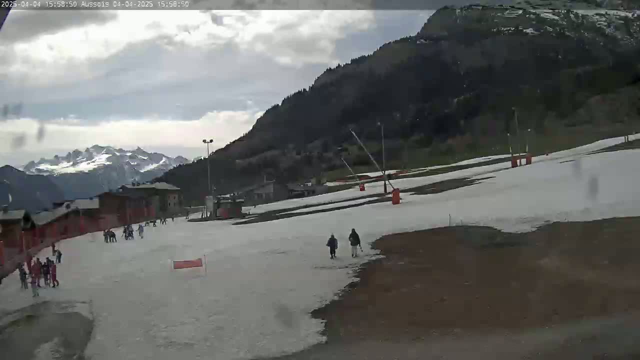 A partly cloudy day shows a snow-covered ski slope with several people scattered across it, a mountain in the background, and several buildings in the distance.