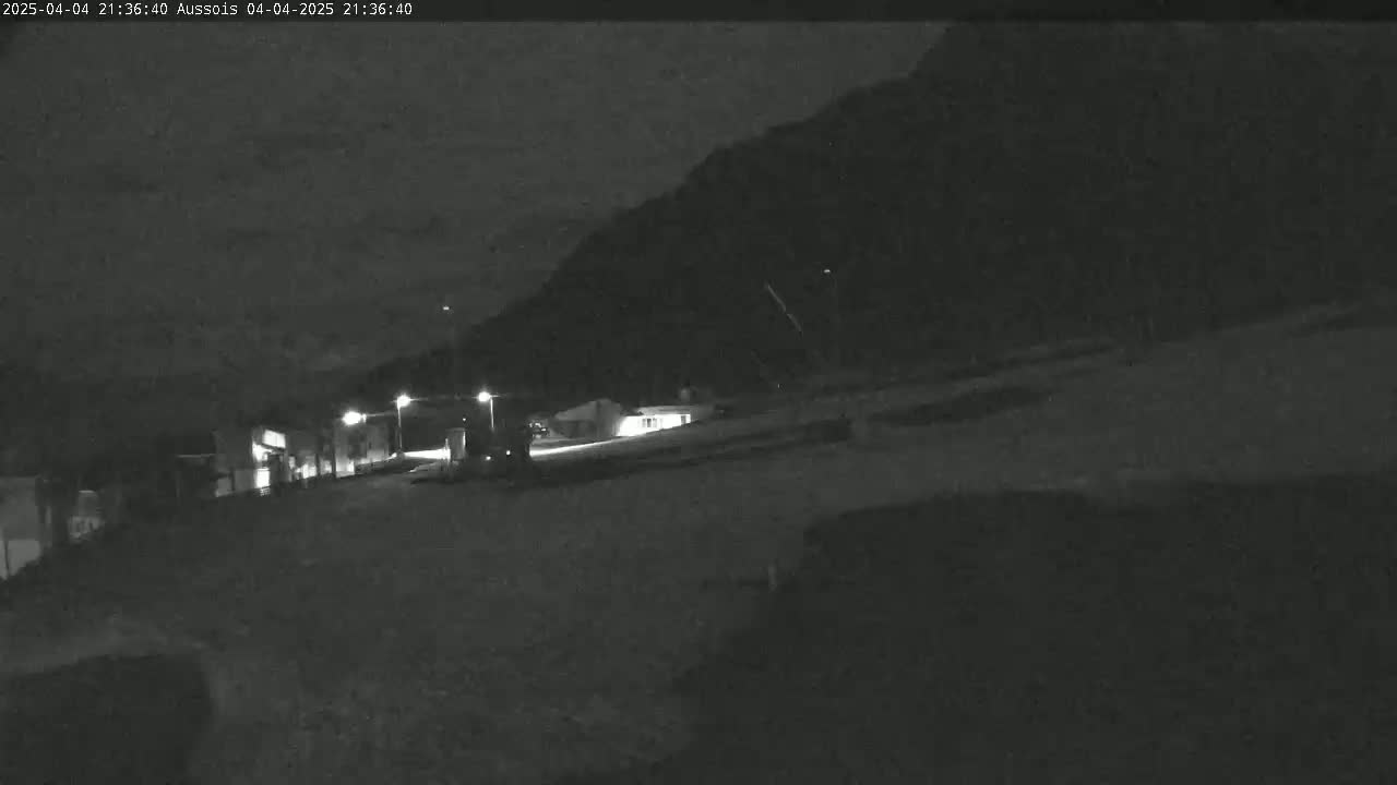 A nighttime, low-light view shows a vehicle driving on a road in front of a dark building against a backdrop of a mountain under a mostly clear sky.