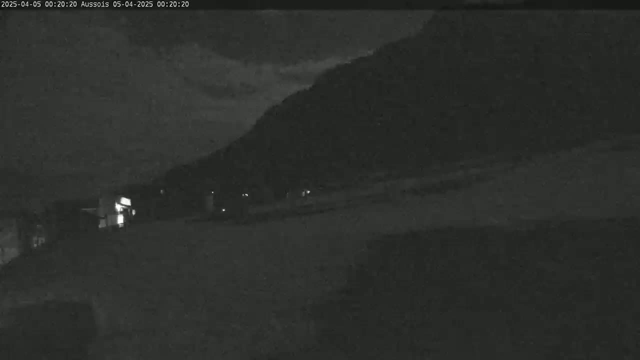 A nighttime, low-light image shows a dark, mostly empty landscape with a hill or mountain in the background under a cloudy sky.