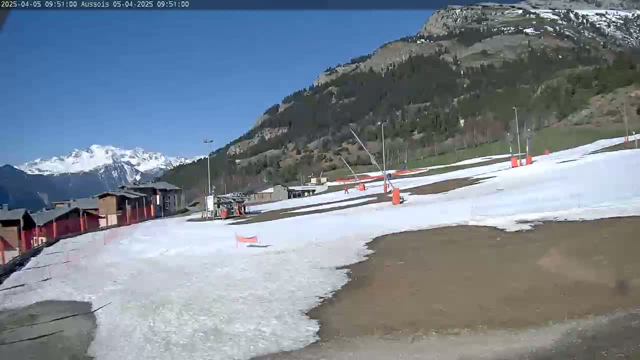 A mostly sunny day reveals a snow-covered ski slope with a few buildings and mountain ranges in the background.