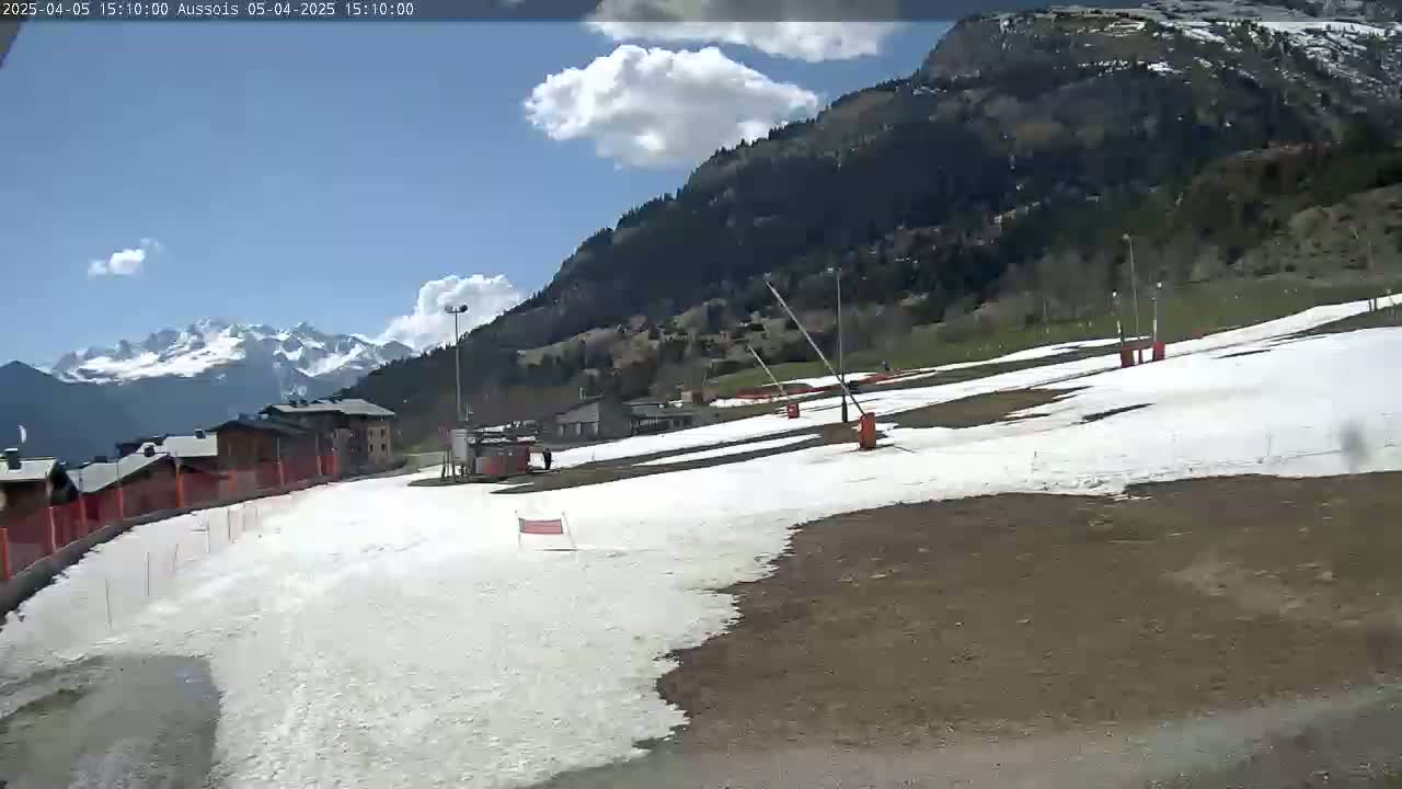 A partly sunny day reveals a mostly snow-covered ski slope in a mountainous area, with buildings and trees visible in the background.