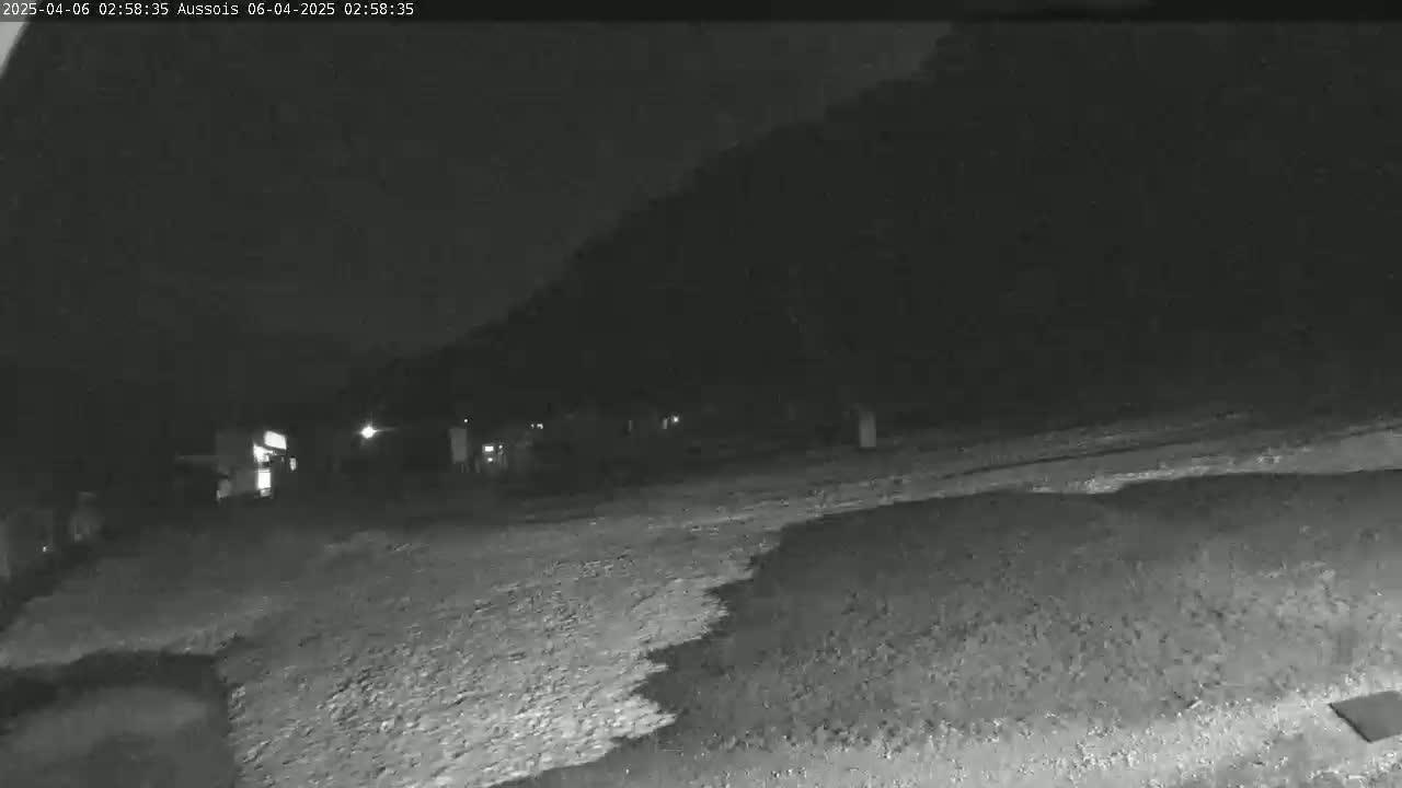 A nighttime, low-light image shows a dark, gravelly area in front of a dimly lit building or structures, with a dark hill or trees rising in the background.