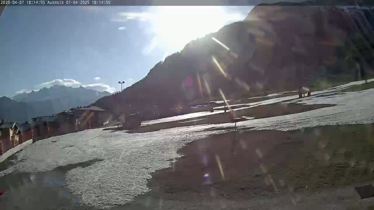 A sunny day in a mountainous area with patchy snow on the ground, showing a few buildings and some people in the distance.