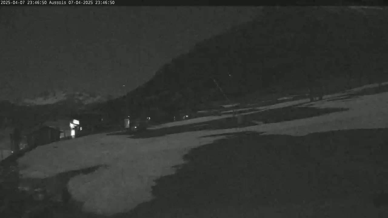 A nighttime, snow-covered landscape shows a few buildings near a dark mountain under a clear sky.