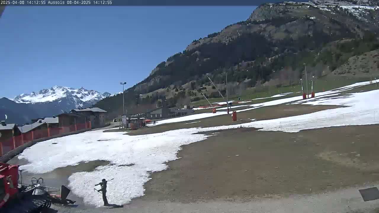 A person tends to a snow-covered area near buildings and a mountainside under a clear blue sky.