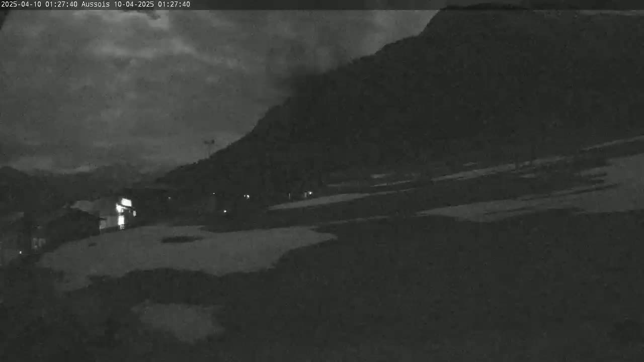 A nighttime, low-light view of a mountainside with some snow patches, a few buildings, and a cloudy sky.