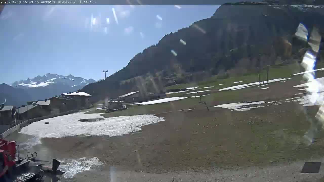 A sunny day in a mountainous region shows a snow-dusted field, a few buildings, and a snowcat in the foreground.