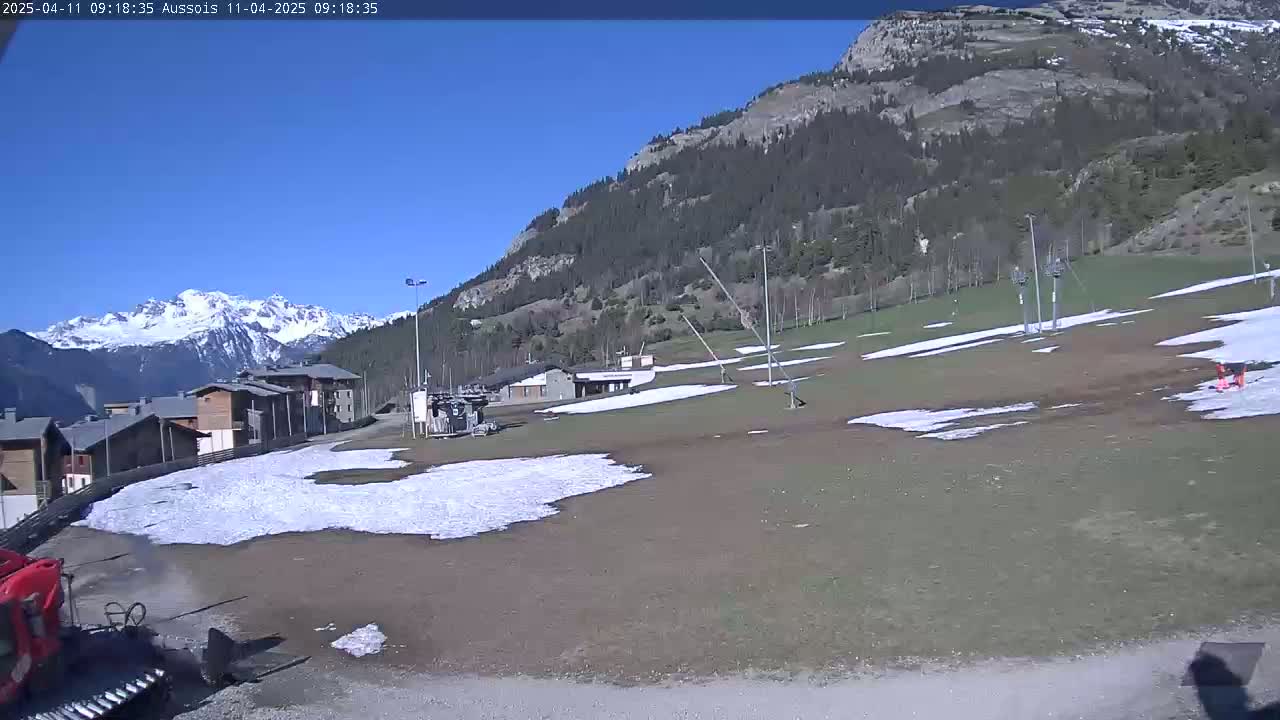 A mostly clear, sunny day reveals a ski resort with patches of melting snow on the ground, a mountain range in the background, and a few buildings.