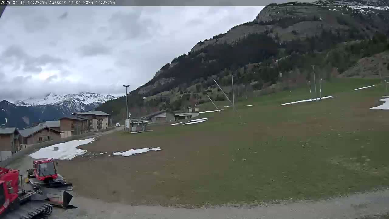 A mostly cloudy day reveals a grassy field with patches of snow, a red snowplow, several buildings, and a mountain range in the background.
