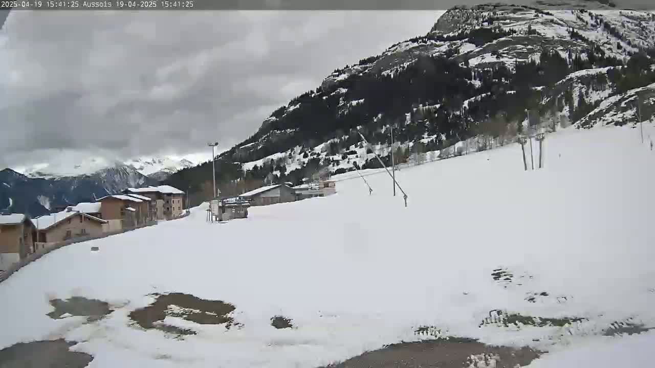 A snow-covered landscape with a few buildings and ski lifts under a cloudy sky.