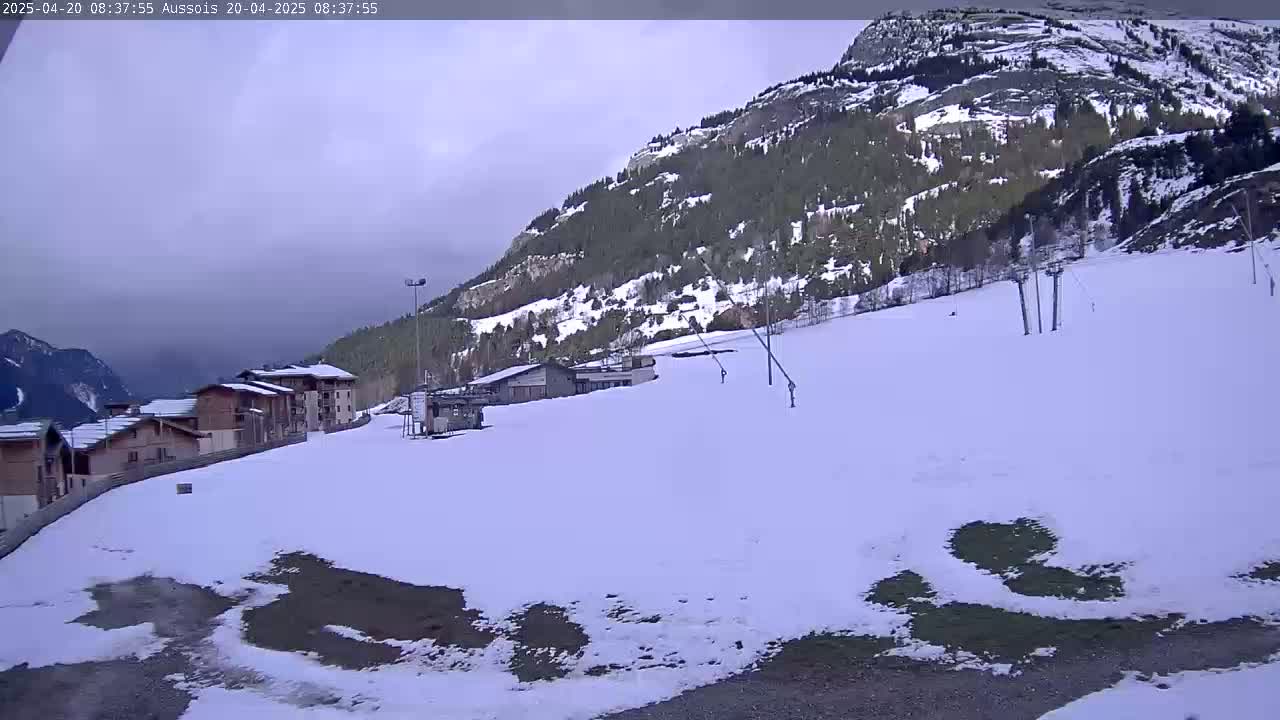 A snow-covered ski slope with buildings and ski lifts sits below a mountain under an overcast sky.
