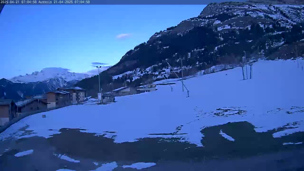 A snow-covered slope in a mountainous region at dawn, with a small village and ski lifts visible in the foreground, and snow-capped mountains in the background.