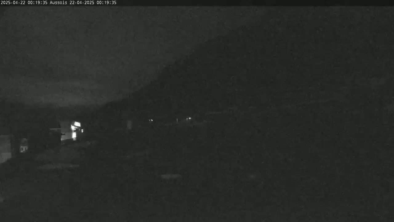 A nighttime, low-light view shows a dark landscape with a hill in the background and a few faint lights visible in the distance.