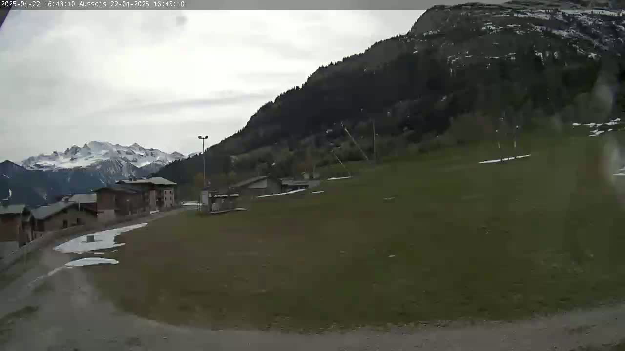 A grassy field with patches of snow and a few buildings sits below a mountain under an overcast sky.